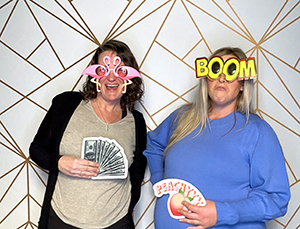 Two women posing with playful photo booth props, including flamingo glasses, a 'BOOM' sign, fake money, and a peach emoji prop, in front of a geometric gold and white backdrop.