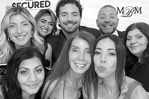 Group photo of eight smiling individuals at an event, with logos reading 'SECURED' and 'M&M' in the background posed for a photo booth.