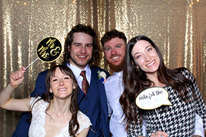 Four smiling individuals posing in a photo booth with a gold sequin backdrop, holding playful props including a 'Mr. & Mrs.' sign and a 'Hello for the Memories' speech bubble