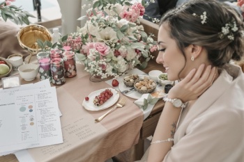 A bride smiling at a beautifully arranged wedding planning table, featuring floral arrangements, dessert plates, and a wedding guide booklet.