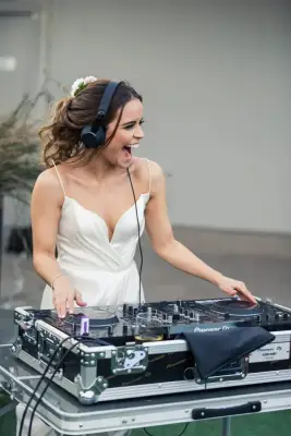 A joyful bride wearing a white dress and headphones, mixing music on a DJ setup at her wedding reception