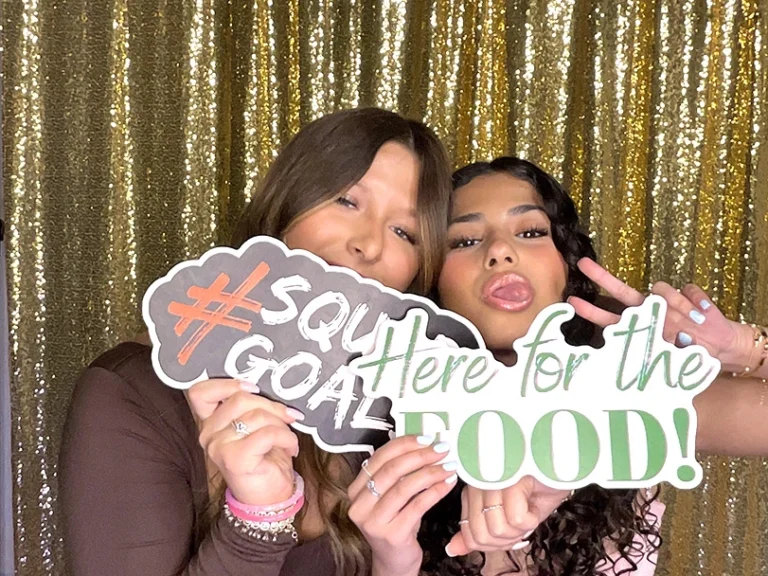 2 girls using a photo booth holding signs that say #squad goals and Here for the food! they're standing in front of a gold sequin backdrop