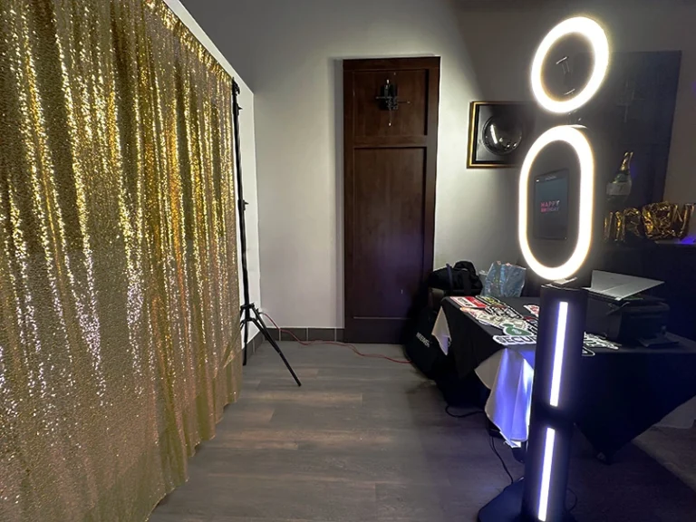 A gold sequined photo booth backdrop with a black table in front of it. There is a white sign on the table with photo props